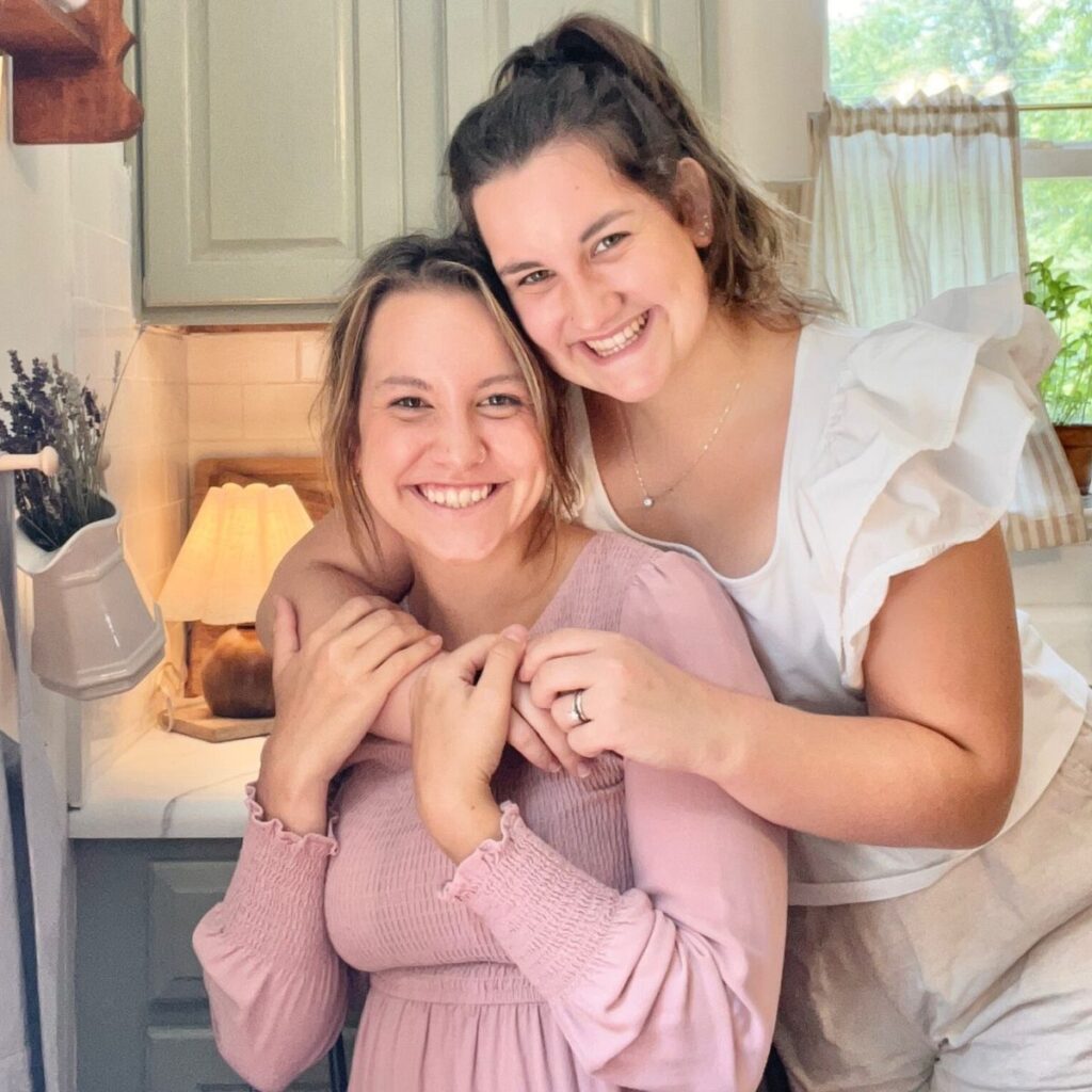A picture of two girls smiling while hugging one another. One girl is wearing a pink, long sleeve dress while the other girl has on a white t shirt. They both have dark hair.