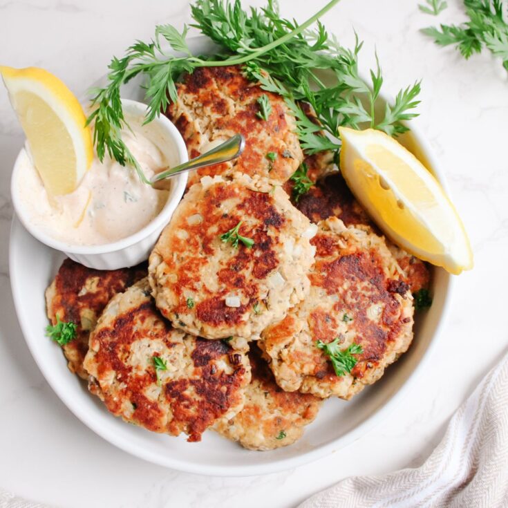 A plate of delicious salmon patties garnished with fresh parsley and lemon slices served alongside salmon sauce.