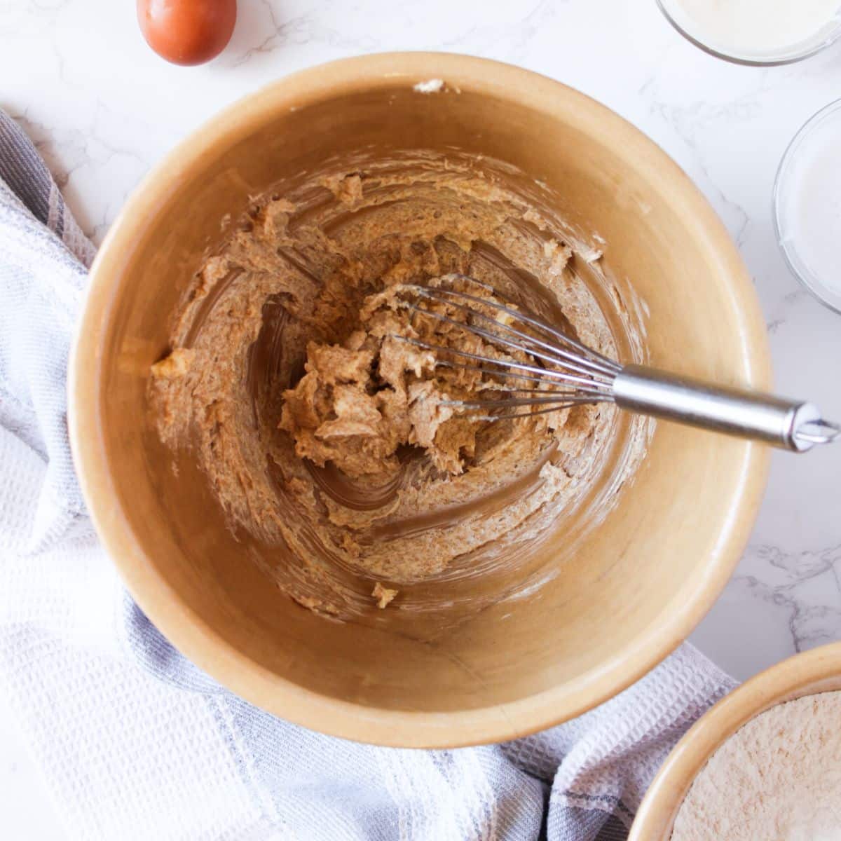 Beat together the butter and sugars with a whisk. In this brown mixing bowl there's brown sugar and butter smoothly mixed together with a silver whisk.