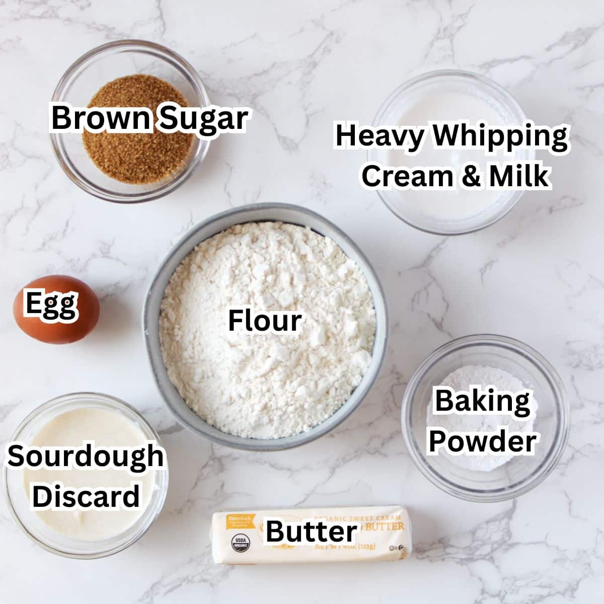 The ingredients for sourdough strawberry shortcake in small bowls-brown sugar, heavy whipping cream and milk, flour, and egg, baking powder, sourdough discard, and butter.