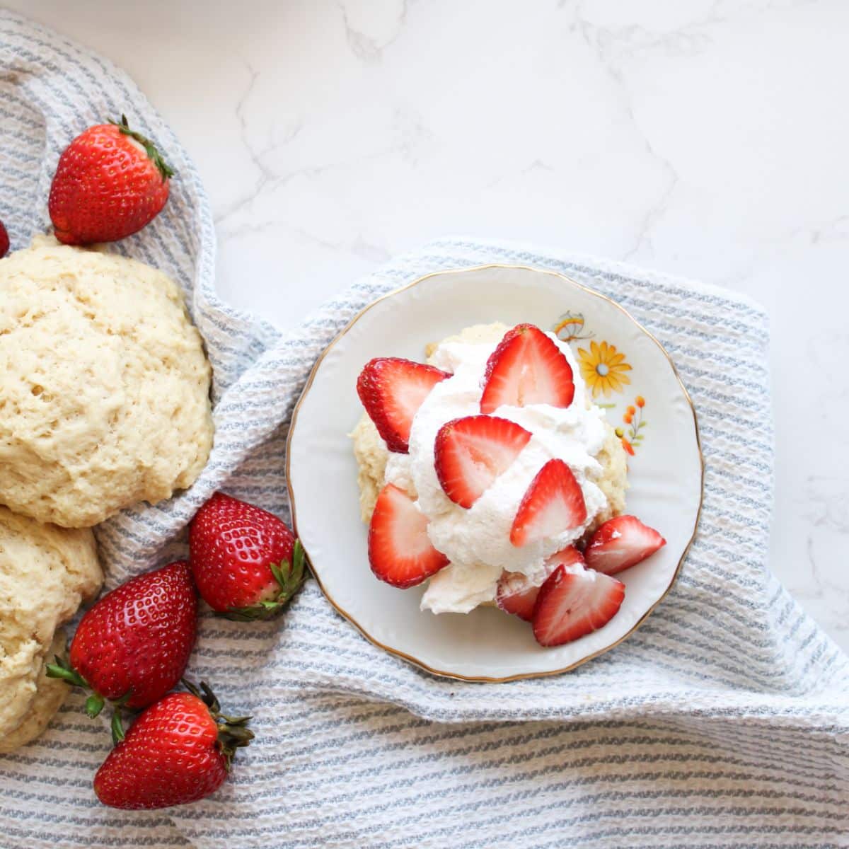 A single serving of sourdough strawberry shortcake on a small plate with sourdough shortcakes and fresh strawberries off to the side.