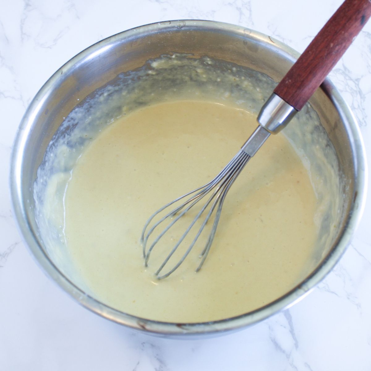 Sourdough crepe batter in a silver mixing bowl with a silver whisk that has a wooden handle.