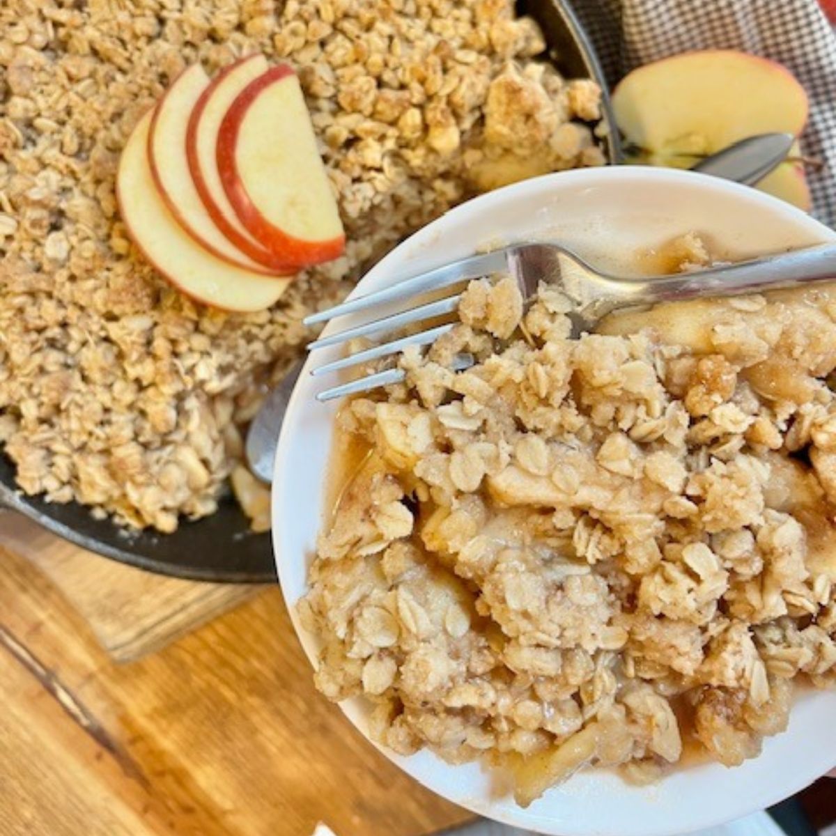 A white plate with apple crisp on it and a silver fork, too. In the background there's a cast iron skillet with apple crisp topped with fresh apple slices.