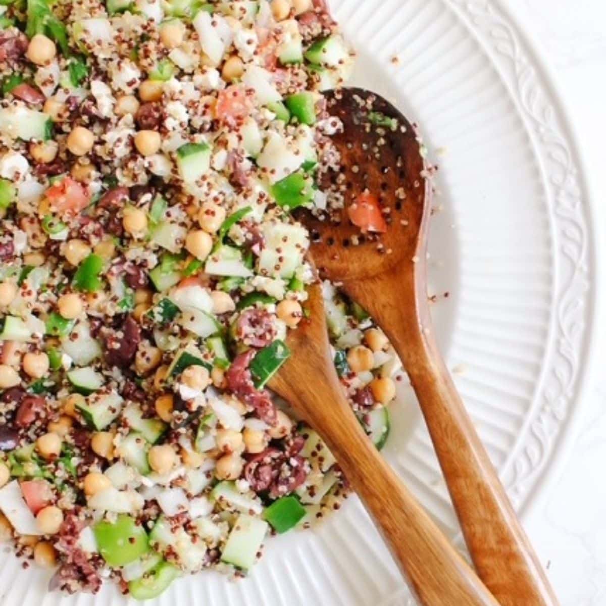 A white bowl filled with quinoa cucumber tomato salad and wooden salad serving utensils.