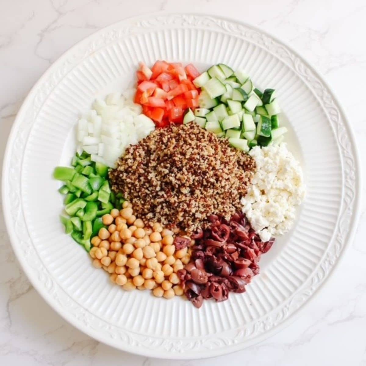 The ingredients you'll need for quinoa cucumber tomato salad in a large white bowl-chick peas, kalamata olives, feta, white onion. green pepper, red tomato, cucumber, and quinoa.