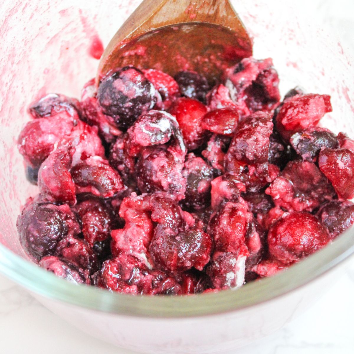 A glass bowl filled with cherries being mixed by a wooden spoon.