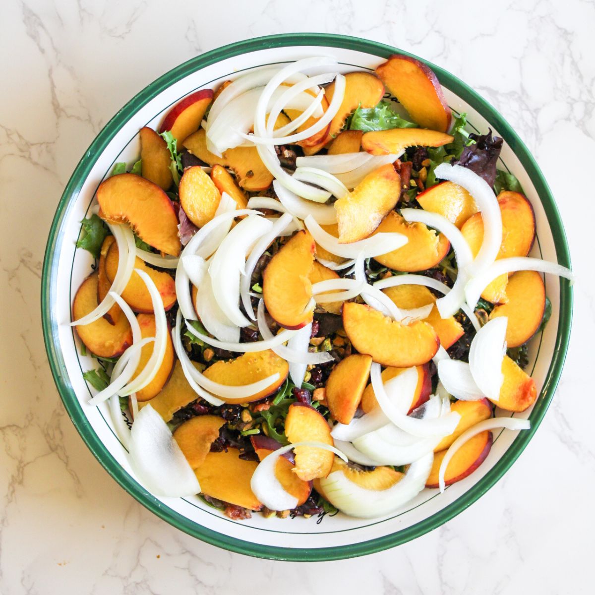 A white bowl that has a green rim filled with a goat cheese peach salad-fresh greens, peach slices, white onion sliced, pistachios and cranberries.