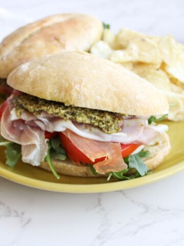 A close up of a pesto panini filled with pesto, prosciutto, tomato slices and fresh arugula, sitting on a yellow plate.