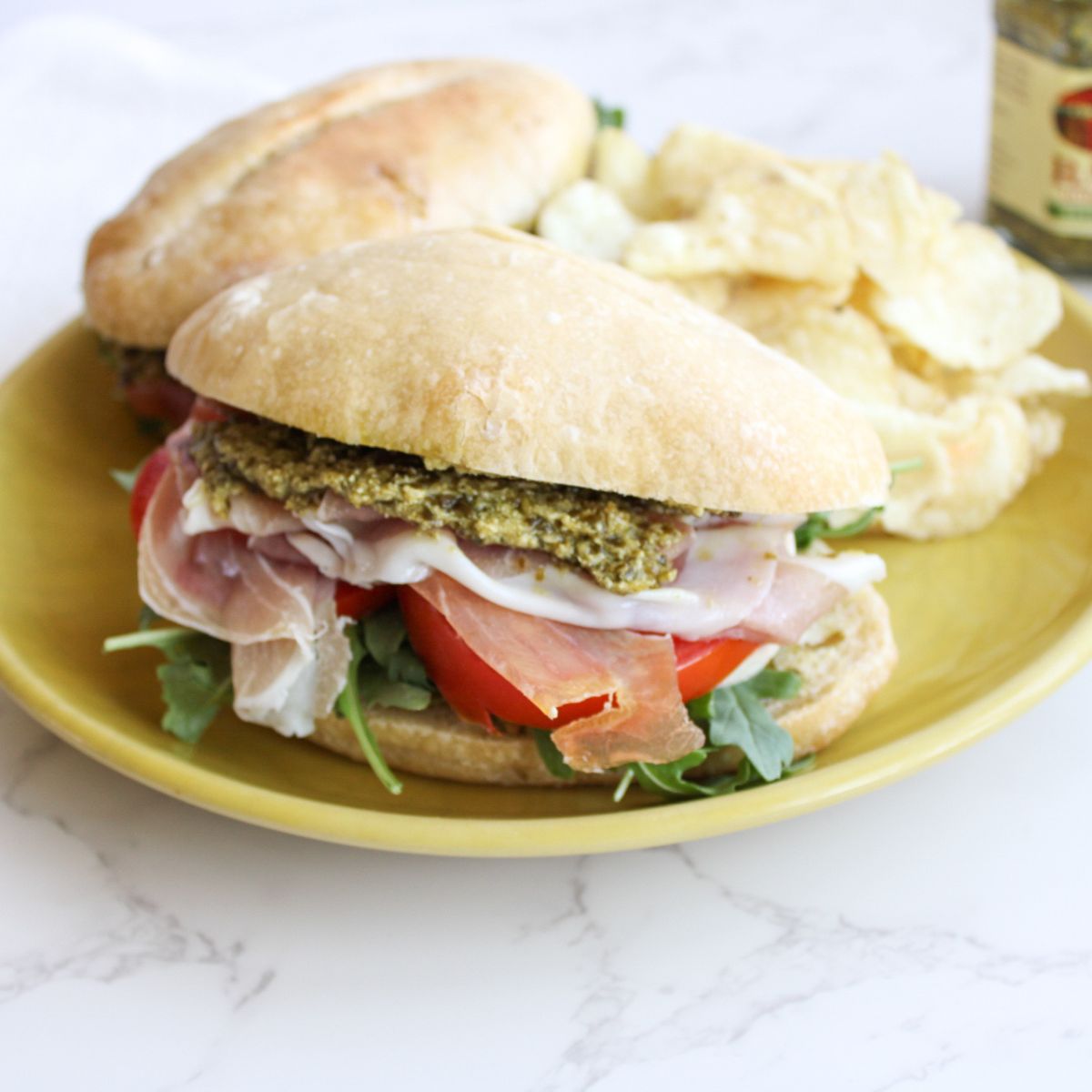 A close up of a pesto panini filled with pesto, prosciutto, tomato slices and fresh arugula, sitting on a yellow plate.