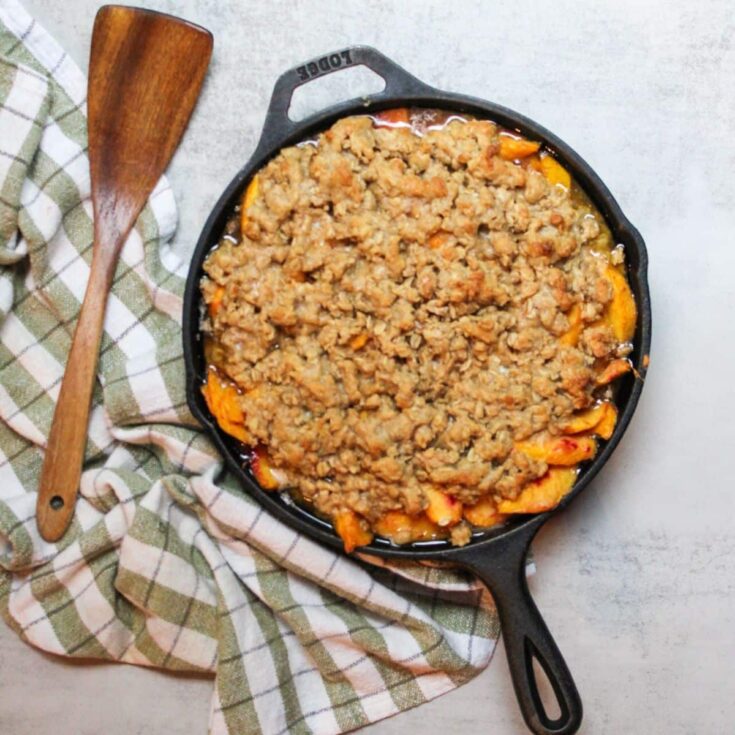 Peach crisp in a cast iron skillet with a wooden serving spoon.
