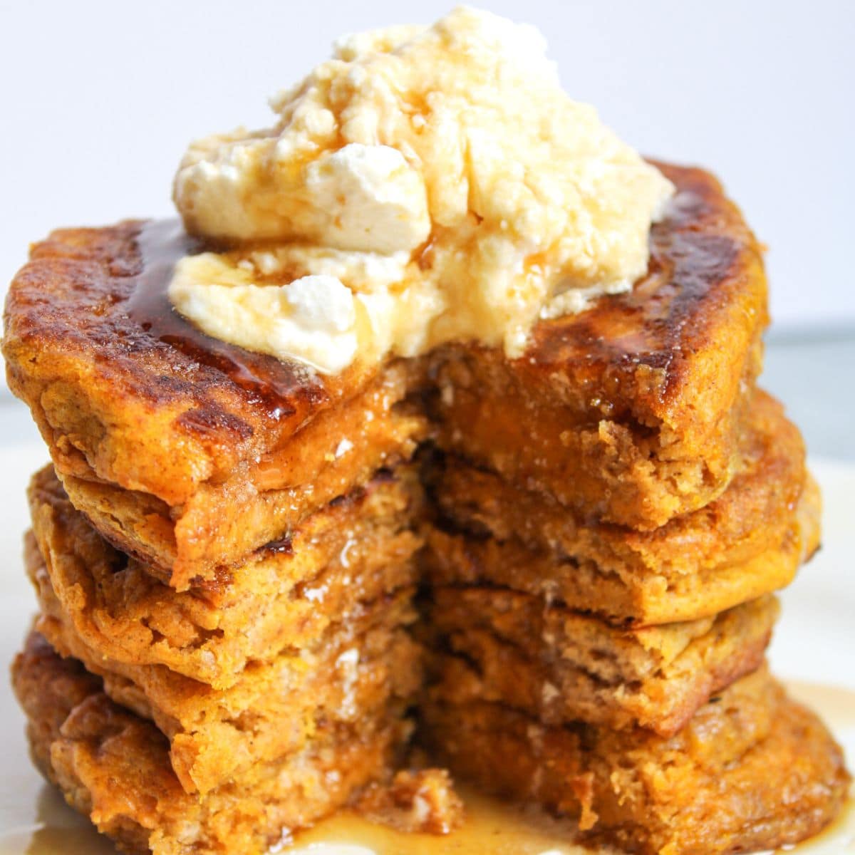 A stack of sourdough pumpkin pancakes with a slice taken out of them, sitting on a white plate and topped with whipped cream and maple syrup.
