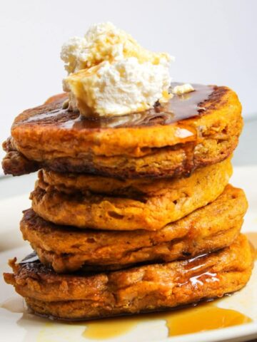 A stack of orange sourdough pumpkin pancakes sitting on a white plate and topped with whipped cream and maple syrup.