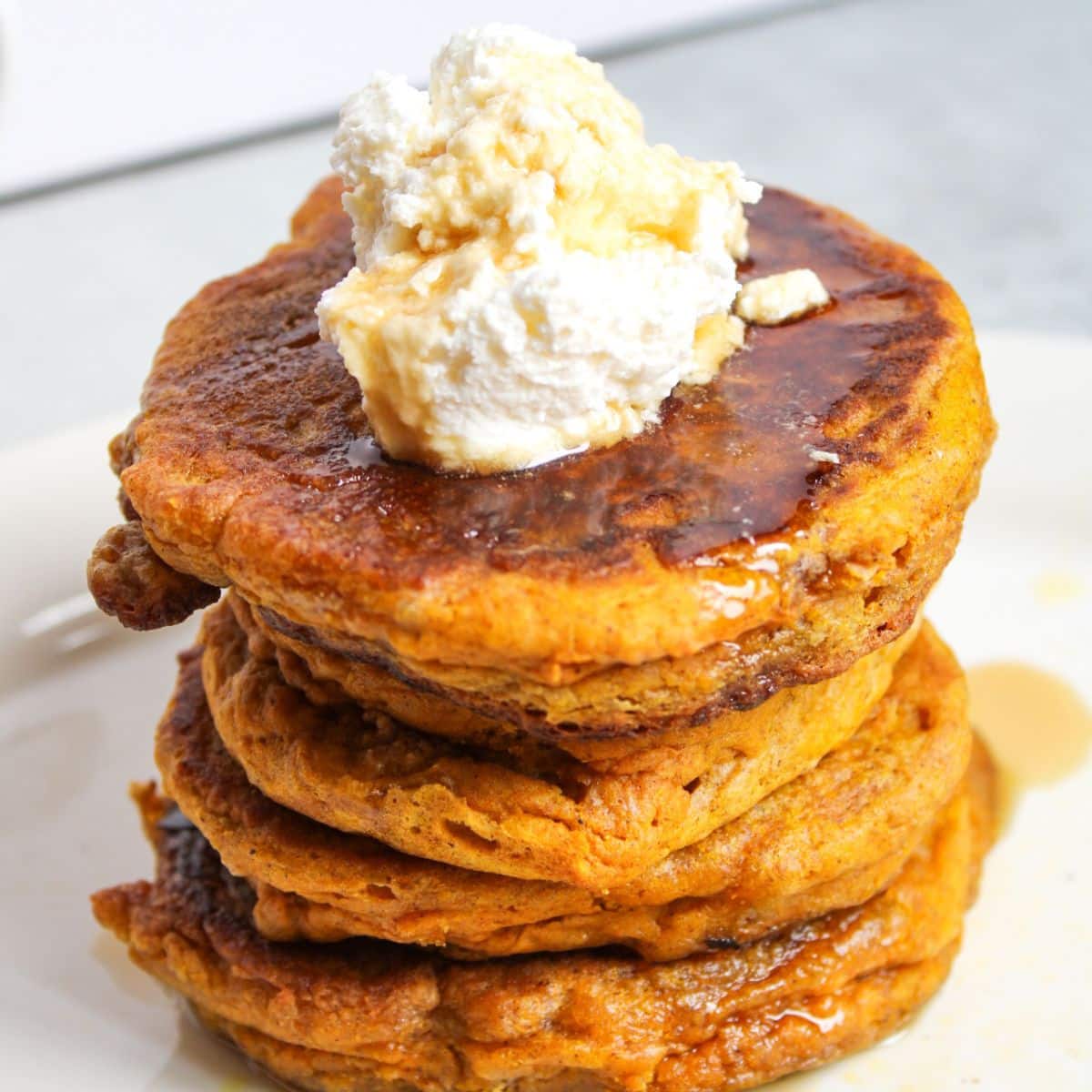 A stack of sourdough pumpkin pancakes sitting on a white plate, topped with whipped cream and maple syrup.