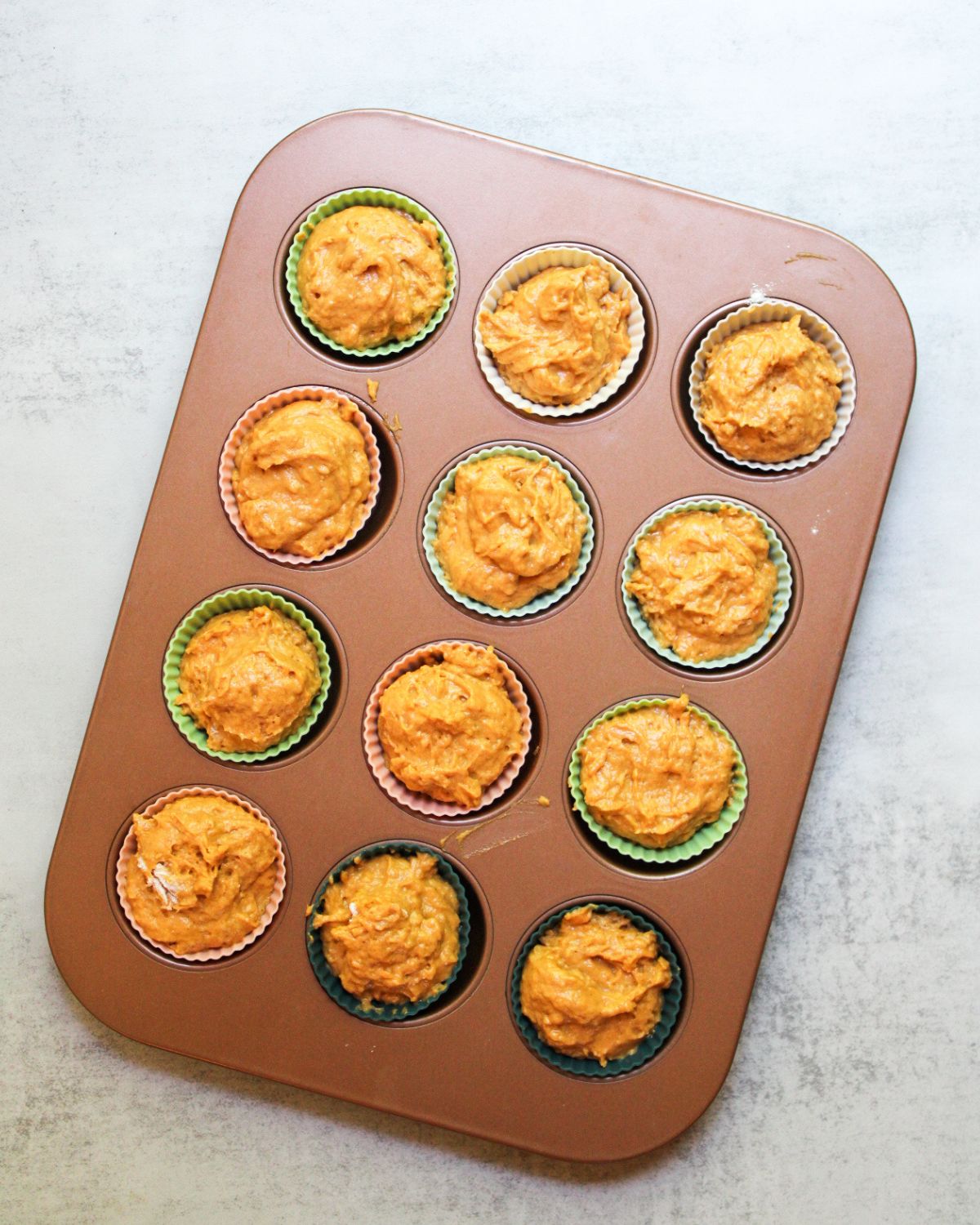 A 12 count muffin tin lined with silicone muffin liners and filled with bright orange sourdough pumpkin muffin batter.