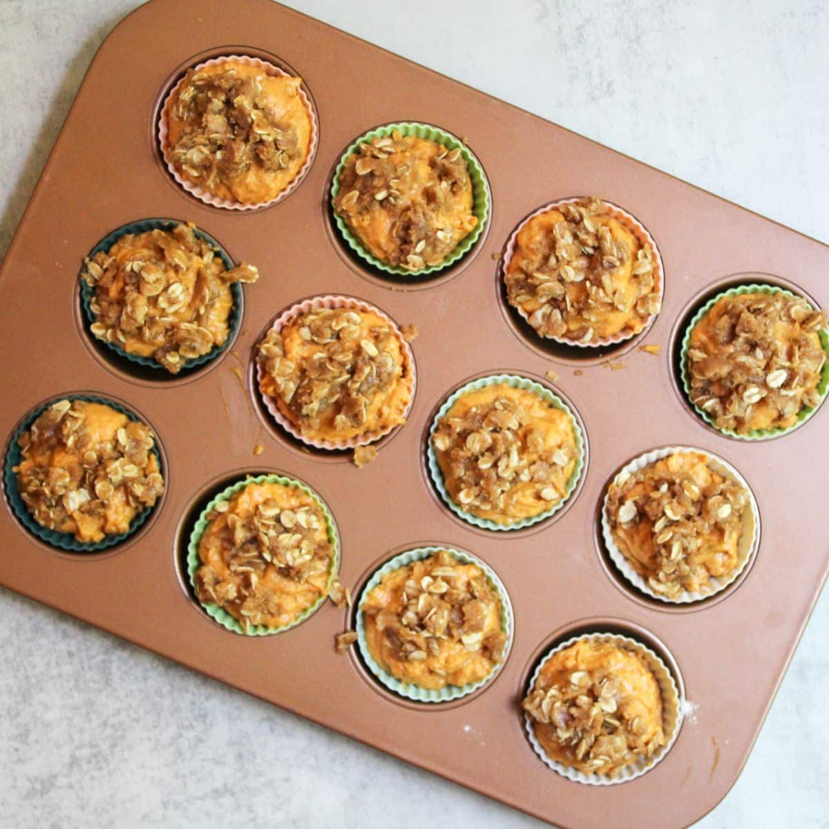 A copper muffin tin filled with silicone muffin liners and each liner has sourdough pumpkin muffin batter. Each pumpkin muffin is topped with a brown sugar oat crumble.
