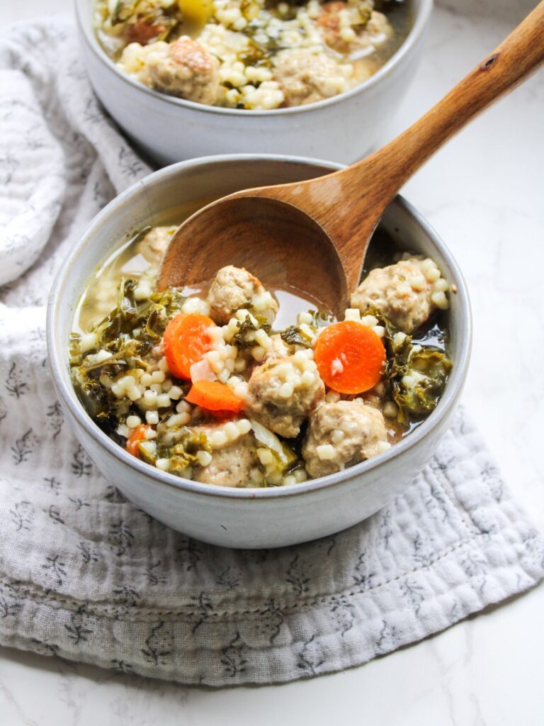 A bowl of Italian wedding soup, on a gray floral napkin with a wooden ladle in the bowl, too.