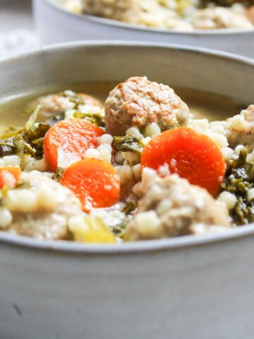 A gray bowl filled with Italian wedding soup that has meatballs, sliced carrots, kale, chicken broth and pasta.
