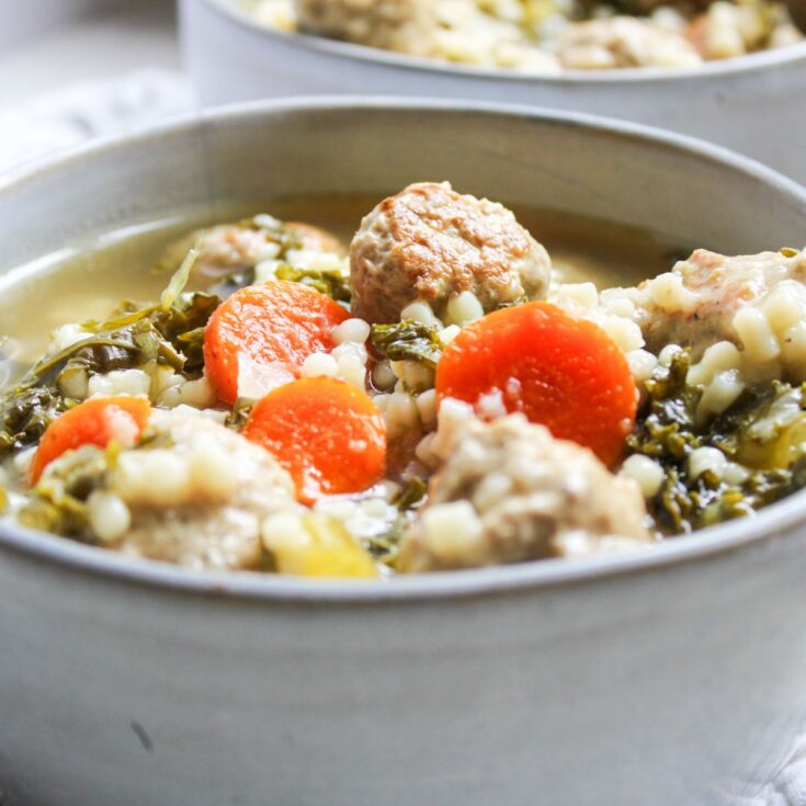 A gray bowl filled with Italian wedding soup that has meatballs, sliced carrots, kale, chicken broth and pasta.