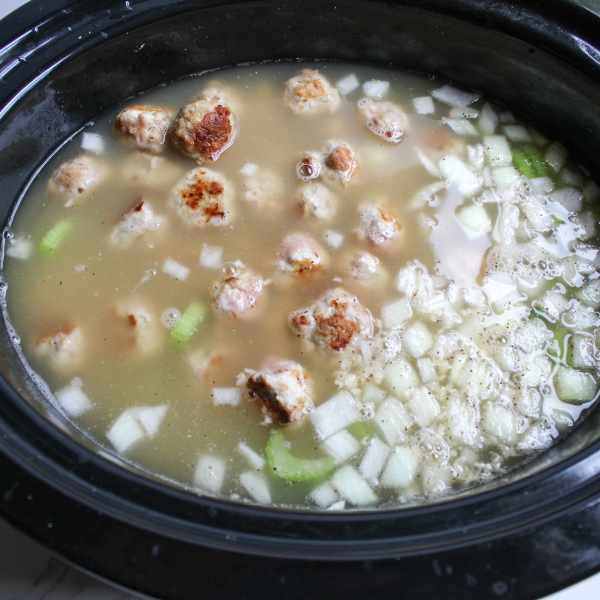 A black crockpot filled with meatballs, diced onion, chopped celery, and chicken broth.