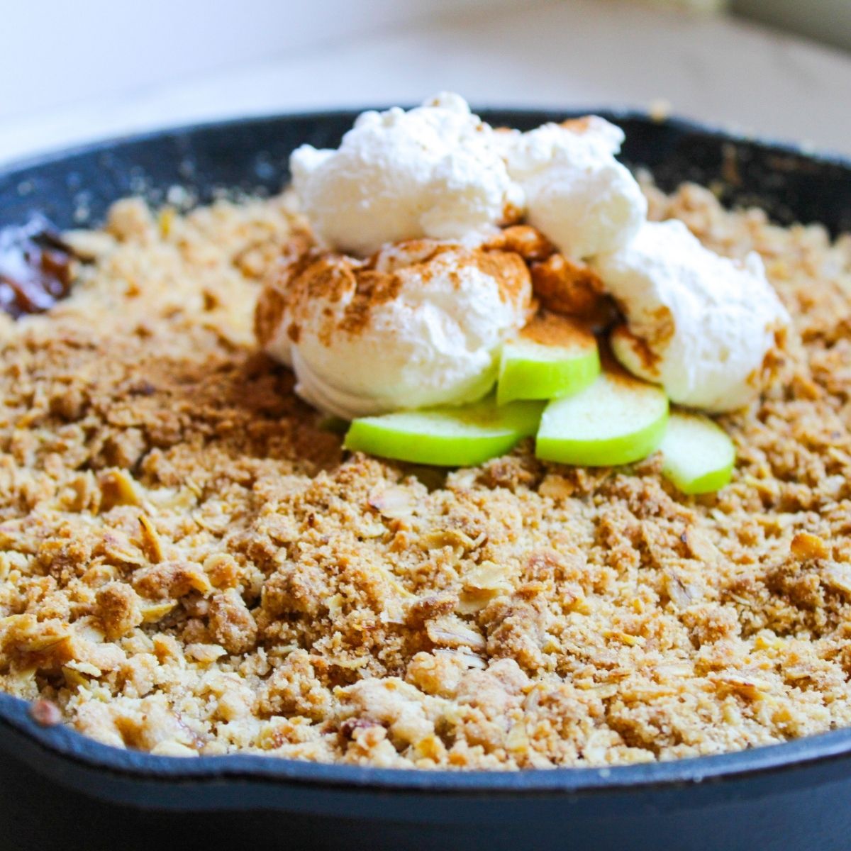 A skillet of apple crisp with oatmeal topping, topped with green apple slices and whipped cream.